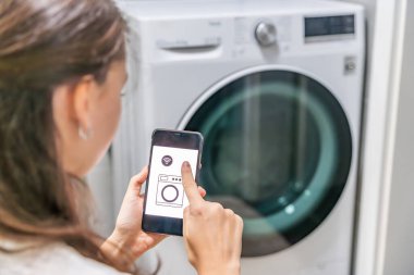 Genç bir kadın cep telefonu kontrolünü kablosuz ağ ve uygulamayla evde çamaşır makinesi olarak kullanıyor. Homesmart ve yeni teknoloji konsepti. Yüksek kalite fotoğraf