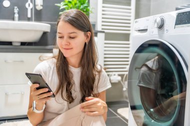 Genç bir kadın cep telefonu kontrolünü kablosuz ağ ve uygulamayla evde çamaşır makinesi olarak kullanıyor. Homesmart ve yeni teknoloji konsepti. Yüksek kalite fotoğraf