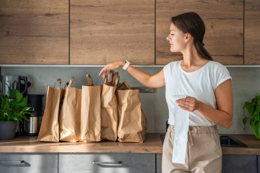 Mutfakta içi yiyecek dolu kağıt torbaların yanındaki dükkandan alınmış fişi olan genç bir kadın. Yiyecekler internetten sipariş edildi ve kuryeyle teslim edildi. Yüksek kalite fotoğraf