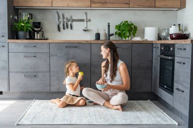 Anne ve kızı mutfak zemininde oturuyor, yemek yiyor ya da kahvaltı edip oynuyorlar. Aile bağı. Yüksek kalite fotoğraf
