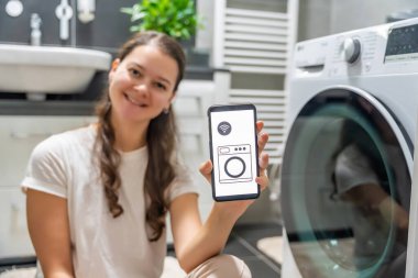 Genç bir kadın cep telefonu kontrolünü kablosuz ağ ve uygulamayla evde çamaşır makinesi olarak kullanıyor. Homesmart ve yeni teknoloji konsepti. Yüksek kalite fotoğraf