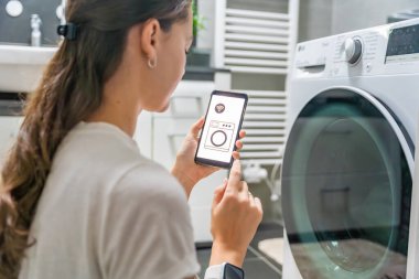 Genç bir kadın cep telefonu kontrolünü kablosuz ağ ve uygulamayla evde çamaşır makinesi olarak kullanıyor. Homesmart ve yeni teknoloji konsepti. Yüksek kalite fotoğraf