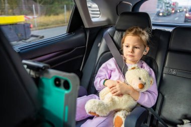 Little girl with teddy bear, tablet and headphones in the car during travel on vacation. Entertainment in the transport for child. High quality photo clipart