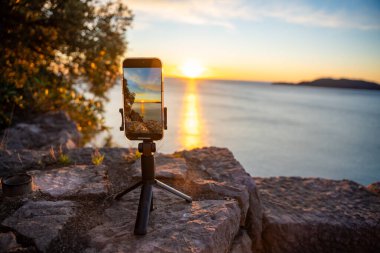 Becici, Karadağ - 1 Ocak 2025: Bir kayanın üzerinde üç ayaklı bir iPhone 'a yakın çekim yaparak, güneşin denizin üzerinde batışının zaman çizelgesini kaydetti. Odaklanma ekranda çarpıcı ufku yakalıyor.. 