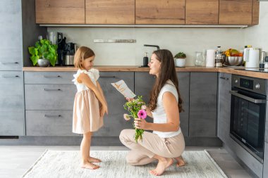 Küçük kız bir buket çiçek veriyor ve ev mutfağında annesine çiziyor. Anneler günü, doğum günü ya da 8. Uluslararası Kadınlar Günü konsepti. Yüksek kalite fotoğraf