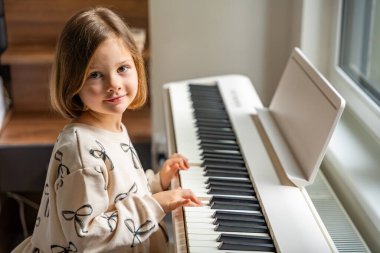 Beyaz piyano sentezleyicili kız için ev müziği dersi. Erken gelişim kavramı: müzik öğrenmek motor becerileri, işitme ve zekayı geliştirir, çocukluktan itibaren yaratıcılığı teşvik eder.. 