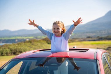 Küçük kız, Kuzey Kıbrıs 'taki dağların arka planına karşı ellerini kaldırarak sevinçle arabanın sunroof' undan sarkıyor. Görüntü özgürlüğü, macerayı ve çocukla seyahat etmenin heyecanını simgeliyor..