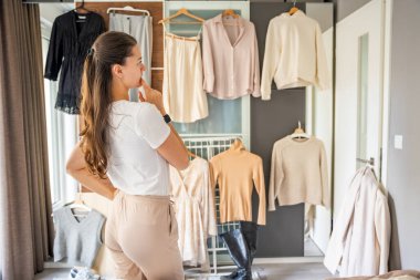 Genç bir kadın elbise dolabının önünde duruyor. Elbiseleri asılmış, ne giyeceğini düşünüyor. Moda seçimleri kişisel stil, ruh hali ve günlük karar verme sürecini yansıtır. 