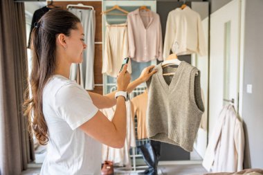 Genç bir kadın, akıllı telefonuyla gardıropta asılı kıyafetleri fotoğraflıyor. Çevrimiçi satış ve ikinci el moda mobil uygulamalar sayesinde gelişiyor, sürdürülebilir alışverişi daha ulaşılabilir hale getiriyor.