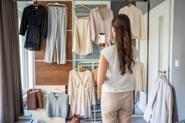 Genç bir kadın, sosyal medya ya da kişisel moda kataloğu için gardıroba asılı giysileri fotoğraflıyor. Dijital platformlar ve çevrimiçi varlık modern moda eğilimlerini şekillendirir. 