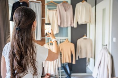 Genç bir kadın, akıllı telefonuyla gardıropta asılı kıyafetleri fotoğraflıyor. Çevrimiçi satış ve ikinci el moda mobil uygulamalar sayesinde gelişiyor, sürdürülebilir alışverişi daha ulaşılabilir hale getiriyor.