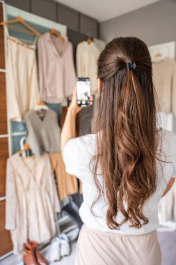 Genç bir kadın, akıllı telefonuyla gardıropta asılı kıyafetleri fotoğraflıyor. Çevrimiçi satış ve ikinci el moda mobil uygulamalar sayesinde gelişiyor, sürdürülebilir alışverişi daha ulaşılabilir hale getiriyor.