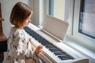 Beyaz piyano sentezleyicili kız için ev müziği dersi. Erken gelişim kavramı: müzik öğrenmek motor becerileri, işitme ve zekayı geliştirir, çocukluktan itibaren yaratıcılığı teşvik eder.. 