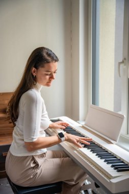 Evde beyaz piyano sentezleyicisiyle oynayan genç bir kadın. Evde müzik dersi kavramı: müzik öğrenmek motor becerileri, işitmeyi ve zekayı geliştirir, yaratıcılığı teşvik eder. Yüksek kalite fotoğraf