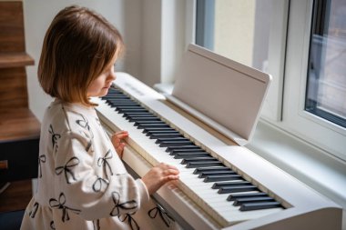 Beyaz piyano sentezleyicili kız için ev müziği dersi. Erken gelişim kavramı: müzik öğrenmek motor becerileri, işitme ve zekayı geliştirir, çocukluktan itibaren yaratıcılığı teşvik eder.. 