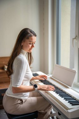 Evde beyaz piyano sentezleyicisiyle oynayan genç bir kadın. Evde müzik dersi kavramı: müzik öğrenmek motor becerileri, işitmeyi ve zekayı geliştirir, yaratıcılığı teşvik eder. Yüksek kalite fotoğraf