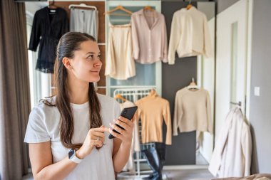 Yatak odasında genç bir kadın, arkasındaki dolapta asılı kıyafetlerle akıllı telefonuna bakıyor. Mobil uygulamalar çevrimiçi satış ve ikinci el moda, sürdürülebilirliği teşvik ve akıllı alışveriş. 