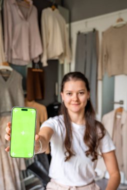 Genç bir kadın kameraya yakın bir akıllı telefon tutuyor, model kullanımı için yeşil bir ekran gösteriyor. Mobil uygulamalar ve dijital aletler çevrimiçi satış ve ikinci el modayı dönüştürür. Yüksek kalite fotoğraf