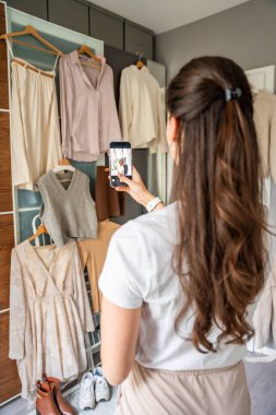Genç bir kadın, sosyal medya ya da kişisel moda kataloğu için gardıroba asılı giysileri fotoğraflıyor. Dijital platformlar ve çevrimiçi varlık modern moda eğilimlerini şekillendirir. 