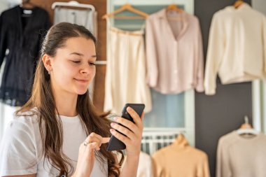 Yatak odasında genç bir kadın, arkasındaki dolapta asılı kıyafetlerle akıllı telefonuna bakıyor. Mobil uygulamalar çevrimiçi satış ve ikinci el moda, sürdürülebilirliği teşvik ve akıllı alışveriş. 