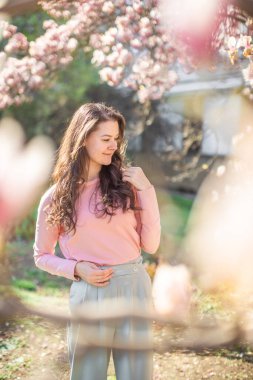 Prag parkında çiçek açan manolya ağaçlarının altında duran zarif genç bir kadın. İlkbaharda moda, doğa ve şehir atmosferlerine modern bir bakış açısı. Yüksek kalite fotoğraf