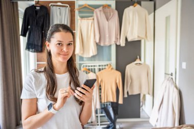 Yatak odasında genç bir kadın, arkasındaki dolapta asılı kıyafetlerle akıllı telefonuna bakıyor. Mobil uygulamalar çevrimiçi satış ve ikinci el moda, sürdürülebilirliği teşvik ve akıllı alışveriş. 