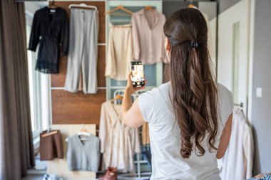 Genç bir kadın, akıllı telefonuyla gardıropta asılı kıyafetleri fotoğraflıyor. Çevrimiçi satış ve ikinci el moda mobil uygulamalar sayesinde gelişiyor, sürdürülebilir alışverişi daha ulaşılabilir hale getiriyor.