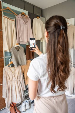 Genç bir kadın, akıllı telefonuyla gardıropta asılı kıyafetleri fotoğraflıyor. Çevrimiçi satış ve ikinci el moda mobil uygulamalar sayesinde gelişiyor, sürdürülebilir alışverişi daha ulaşılabilir hale getiriyor.