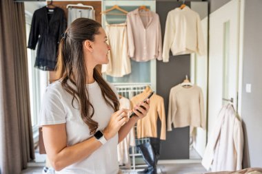 Yatak odasında genç bir kadın, arkasındaki dolapta asılı kıyafetlerle akıllı telefonuna bakıyor. Mobil uygulamalar çevrimiçi satış ve ikinci el moda, sürdürülebilirliği teşvik ve akıllı alışveriş. 