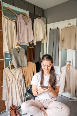 Yatak odasında genç bir kadın, arkasındaki dolapta asılı kıyafetlerle akıllı telefonuna bakıyor. Mobil uygulamalar çevrimiçi satış ve ikinci el moda, sürdürülebilirliği teşvik ve akıllı alışveriş. 