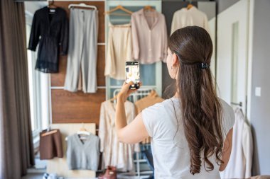 Genç bir kadın, akıllı telefonuyla gardıropta asılı kıyafetleri fotoğraflıyor. Çevrimiçi satış ve ikinci el moda mobil uygulamalar sayesinde gelişiyor, sürdürülebilir alışverişi daha ulaşılabilir hale getiriyor.