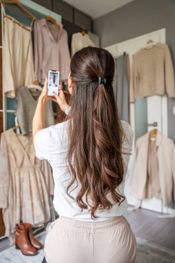 Genç bir kadın, akıllı telefonuyla gardıropta asılı kıyafetleri fotoğraflıyor. Çevrimiçi satış ve ikinci el moda mobil uygulamalar sayesinde gelişiyor, sürdürülebilir alışverişi daha ulaşılabilir hale getiriyor.