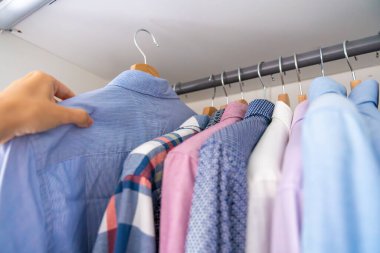 Erkek işçi, ahşap askılarda giysi dolu bir dolaptan gömlek seçiyor. Kıyafet seçimi kavramı, günlük rutin ve kişisel stil. Yüksek kalite fotoğraf