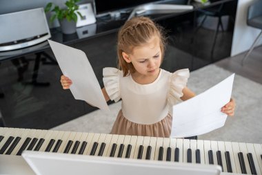 Evde piyano çalışmasından önce nota okuyan küçük bir kız. Konforlu bir aile ortamında müzikal öğrenme ve hazırlık odaklı bir an. Yüksek kalite fotoğraf