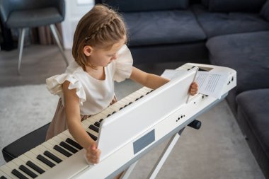 Küçük kız evde oturma odasında piyano çalmaya hazırlanıyor. Konforlu bir aile ortamında müzik çalışmasından önce sakin bir odak ve beklenti anı. Yüksek kalite fotoğraf