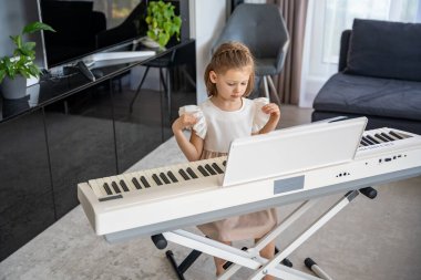 Küçük kız evde oturma odasında piyano çalmaya hazırlanıyor. Konforlu bir aile ortamında müzik çalışmasından önce sakin bir odak ve beklenti anı. Yüksek kalite fotoğraf