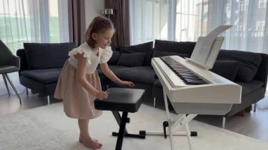 Küçük kız evde oturma odasında piyano çalmaya hazırlanıyor. Konforlu bir aile ortamında müzik çalışmasından önce sakin bir odak ve beklenti anı. Yüksek kalite 4k görüntü