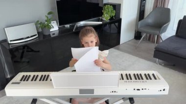 Evde piyano çalışmasından önce nota okuyan küçük bir kız. Konforlu bir aile ortamında müzikal öğrenme ve hazırlık odaklı bir an. Yüksek kalite 4k görüntü