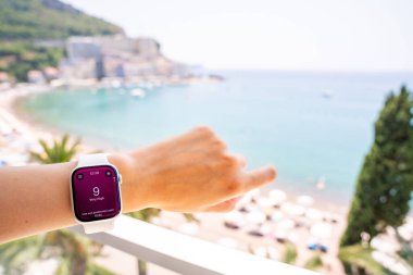Budva, Karadağ - 12 Haziran 2025: Bileklerdeki zeka seviyesi, arka planda plaj, deniz ve güneş bulunan yüksek UV endeksini gösteriyor. Giyilebilir teknoloji yaz tatilinde ultraviyole ışığın izlenmesine yardımcı olur.. 