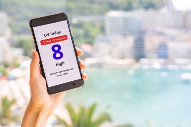 Akıllı telefon arka planda bulanık sahil, deniz ve şehirle birlikte yüksek UV uyarısı gösteriyor. Sıcak güneşli hava ve radyasyon uyarısı. Yüksek kalite fotoğraf