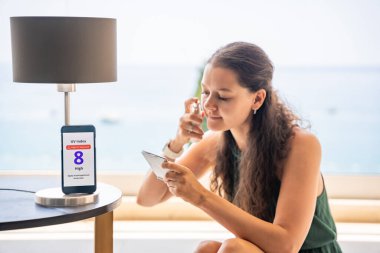 Kadın aynada yüzüne güneş kremi sürerken, odakta yüksek UV uyarısı olan akıllı telefon. Bilinçli cilt bakım rutini güneş güvenlik uyarısından esinlenilmiştir. Yüksek kalite fotoğraf