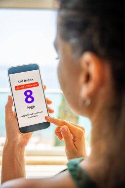 Genç bir kadın yüksek radyasyon uyarısıyla akıllı telefondan ultraviyole veri tabanına bakıyor. Mobil uygulama dışarı çıkmadan önce güneş kremi sürmeyi uyarıyor. Yüksek kalite fotoğraf