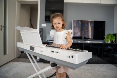 Küçük kız evde oturma odasında piyano çalmaya hazırlanıyor. Konforlu bir aile ortamında müzik çalışmasından önce sakin bir odak ve beklenti anı. Yüksek kalite fotoğraf