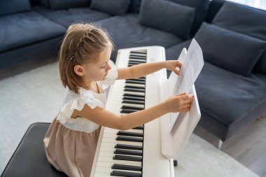Evde piyano çalışmasından önce nota okuyan küçük bir kız. Konforlu bir aile ortamında müzikal öğrenme ve hazırlık odaklı bir an. Yüksek kalite fotoğraf