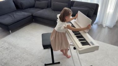 Küçük kız evde oturma odasında piyano çalmaya hazırlanıyor. Konforlu bir aile ortamında müzik çalışmasından önce sakin bir odak ve beklenti anı. Yüksek kalite 4k görüntü