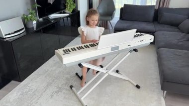 Küçük kız evde oturma odasında piyano çalmaya hazırlanıyor. Konforlu bir aile ortamında müzik çalışmasından önce sakin bir odak ve beklenti anı. Yüksek kalite 4k görüntü