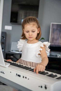 Küçük kız evde oturma odasında piyano çalmaya hazırlanıyor. Konforlu bir aile ortamında müzik çalışmasından önce sakin bir odak ve beklenti anı. Yüksek kalite fotoğraf
