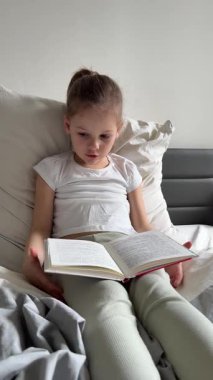 Küçük kız ailesinin yatağında oturuyor ve yatak odasında kitap okuyor. Sessiz zaman kavramı, konsantrasyon ve erken yaşta okuma aşkı. Yüksek kalite 4k görüntü