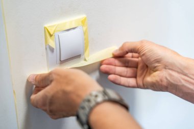 Duvarı beyaza boyamadan önce adam lambanın düğmesini bantla kapatıyor. Ev tadilatı, yüzey koruması ve iç işleri sırasında dikkatli bir hazırlık. Yüksek kalite fotoğraf