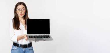 Portrait of cute girl in glasses, student showing laptop screen with amazed face, standing over white background. Copy space